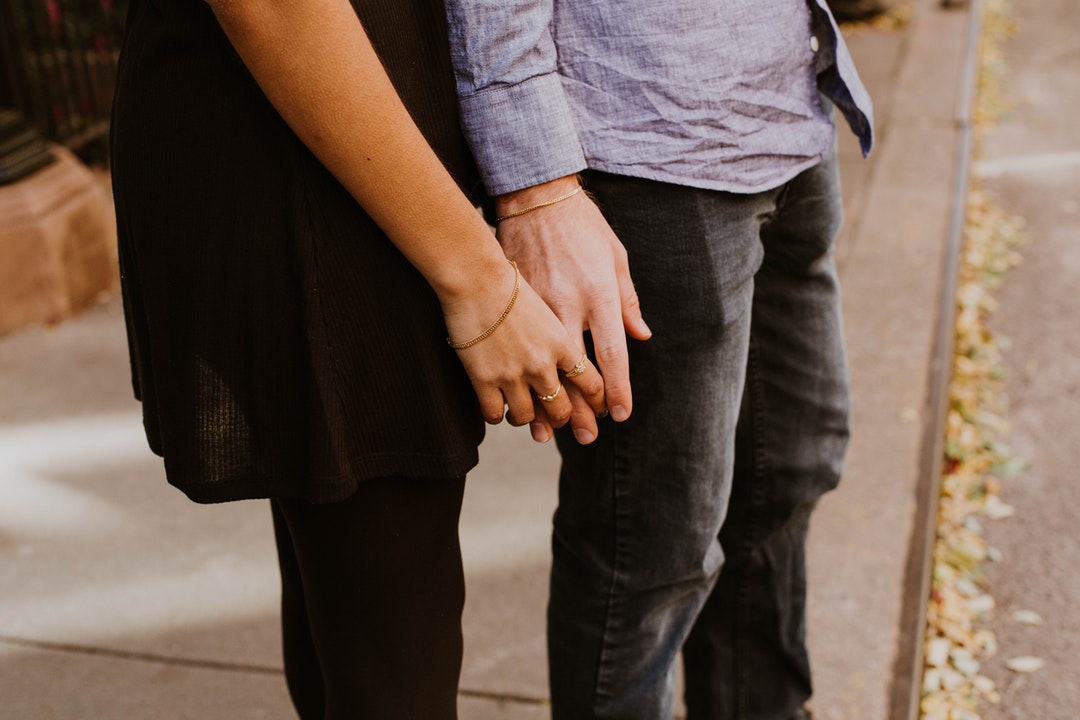 A couple holding hands, standing on a sidewalk. Both are wearing bracelets.