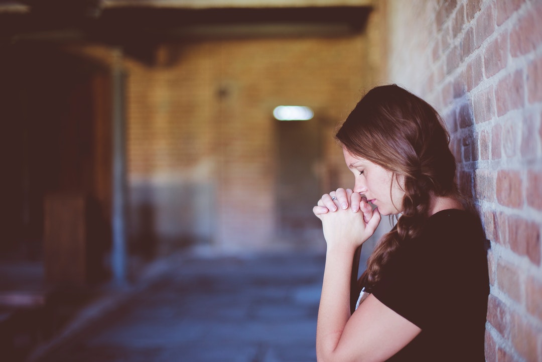 A woman wearing a braid in her hair is leaning against a brick wall, praying