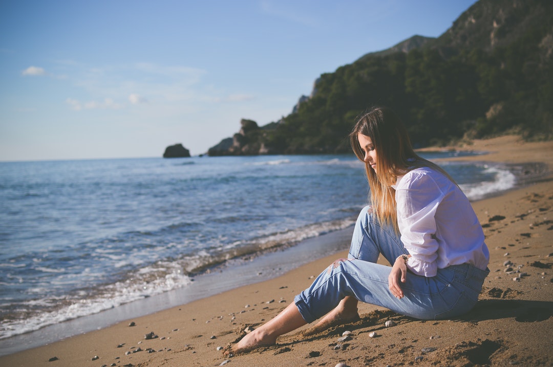 Young woman sitting on a sand beach in white shirt and jeans in Corfu