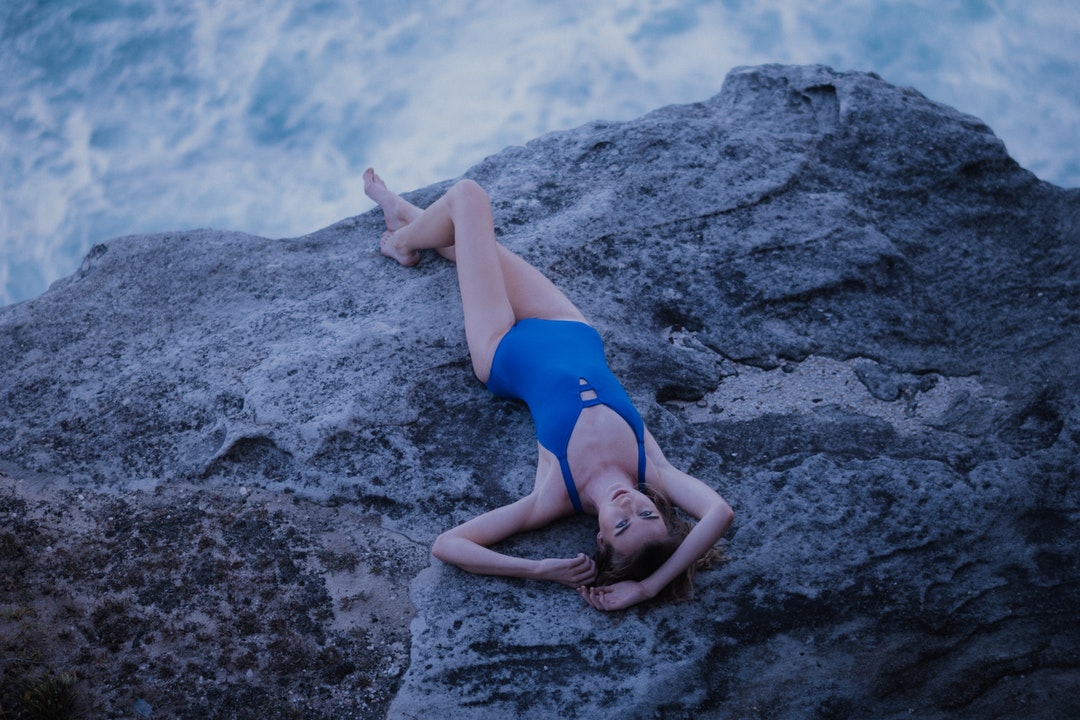 Woman in a blue swimsuit lying down on a rock overlooking the sea at Bondi Beach