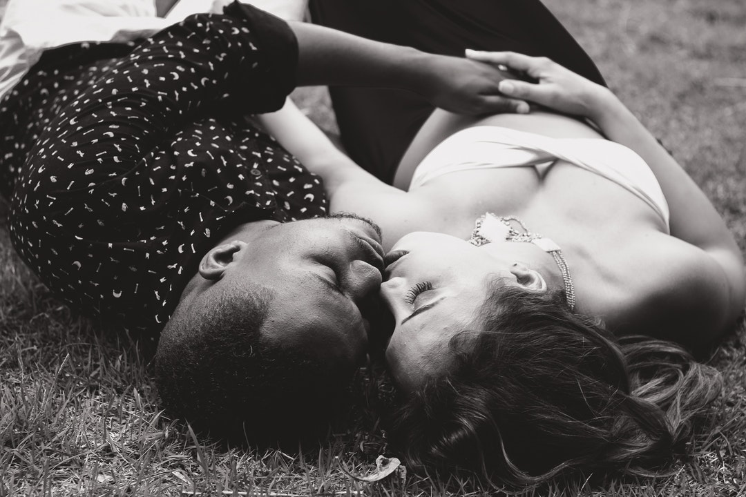 A man and a woman lying down on grass cuddle and kiss in black-and-white