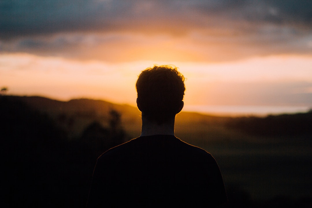 Silhouette of the back of a man's head, looking at the sun in a cloudy, orange sky