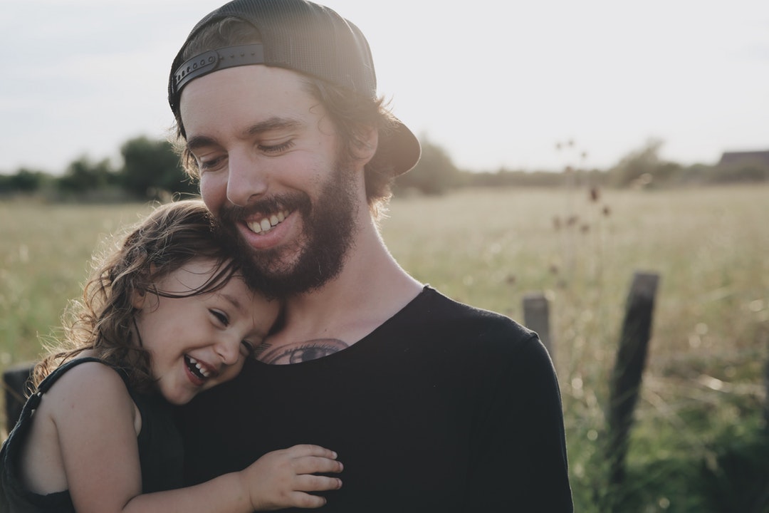Man with a beard, tattoo and black shirt holds a laughing girl in a field in the sun