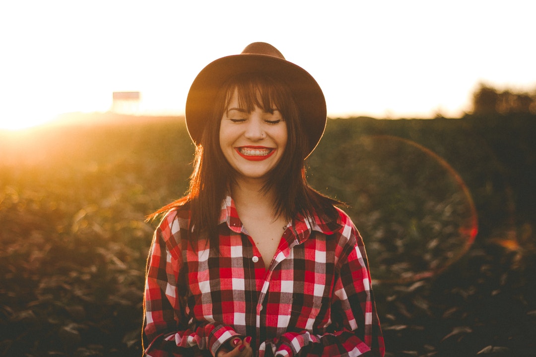 Woman in bright red lipstick, a hat, and a plaid shirt smiling in a sunny field