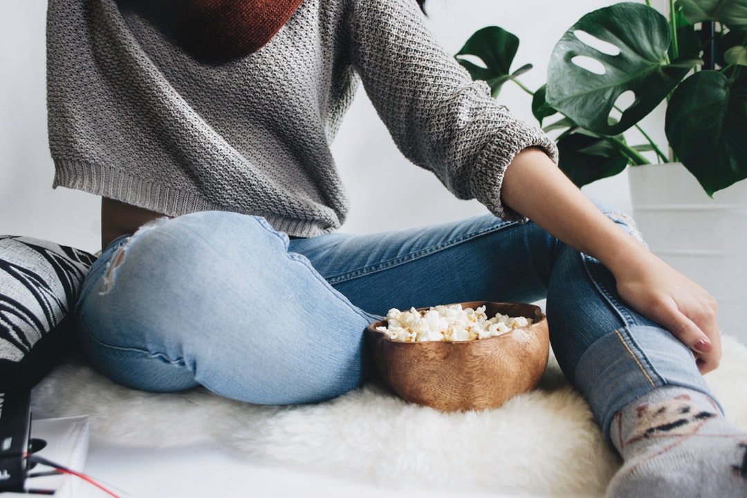 Woman in a cropped gray sweater sits on a furry rug with a bowl of popcorn