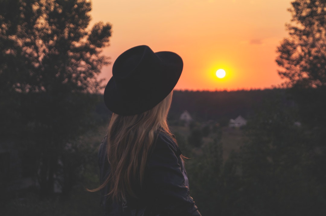 Back view of a blonde woman with a black hat on her head, watching the sunset over trees