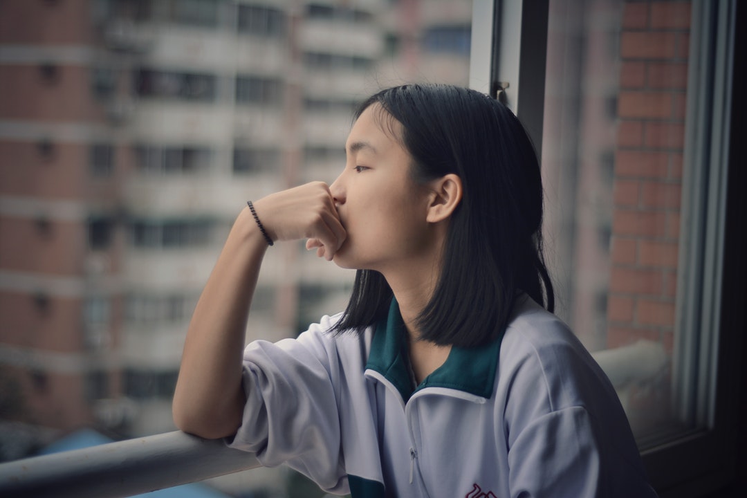 A teenager looks out the window with a hand on mouth and elbows on the railing.