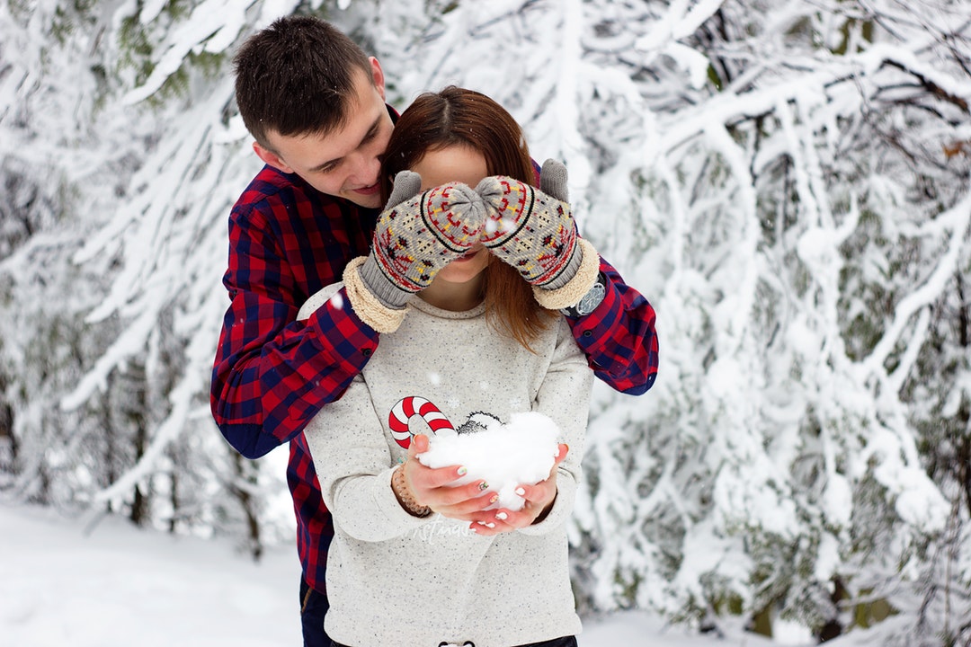 A man stands behind a woman and covers her eyes with his hands in mittens; she is laughing and holding a pile of snow in her hands