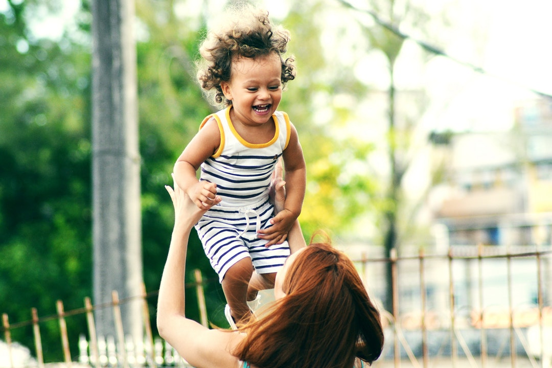 A young girl with curly hair wearing a striped outfit laughing as her brunette mom throws her in the air, Salvador