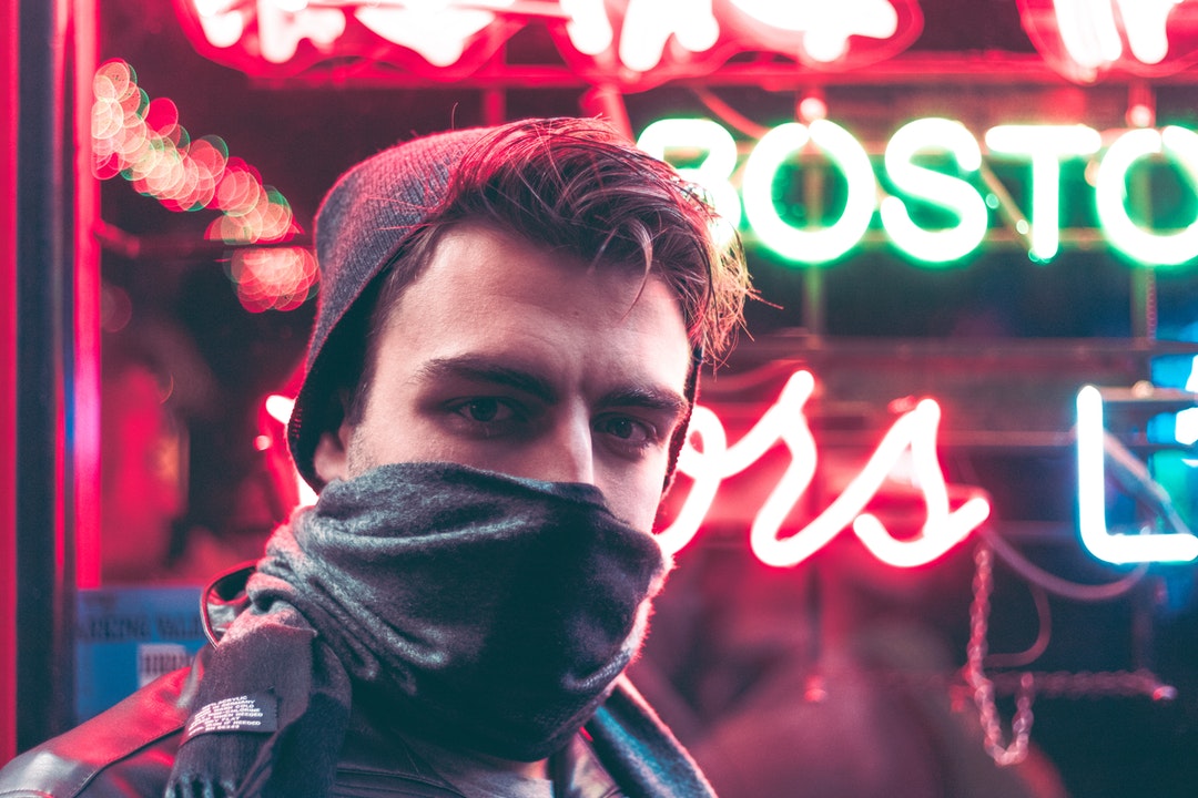 A man wearing a hat, covering his face with a scarf and standing in front of a neon sign in Boston