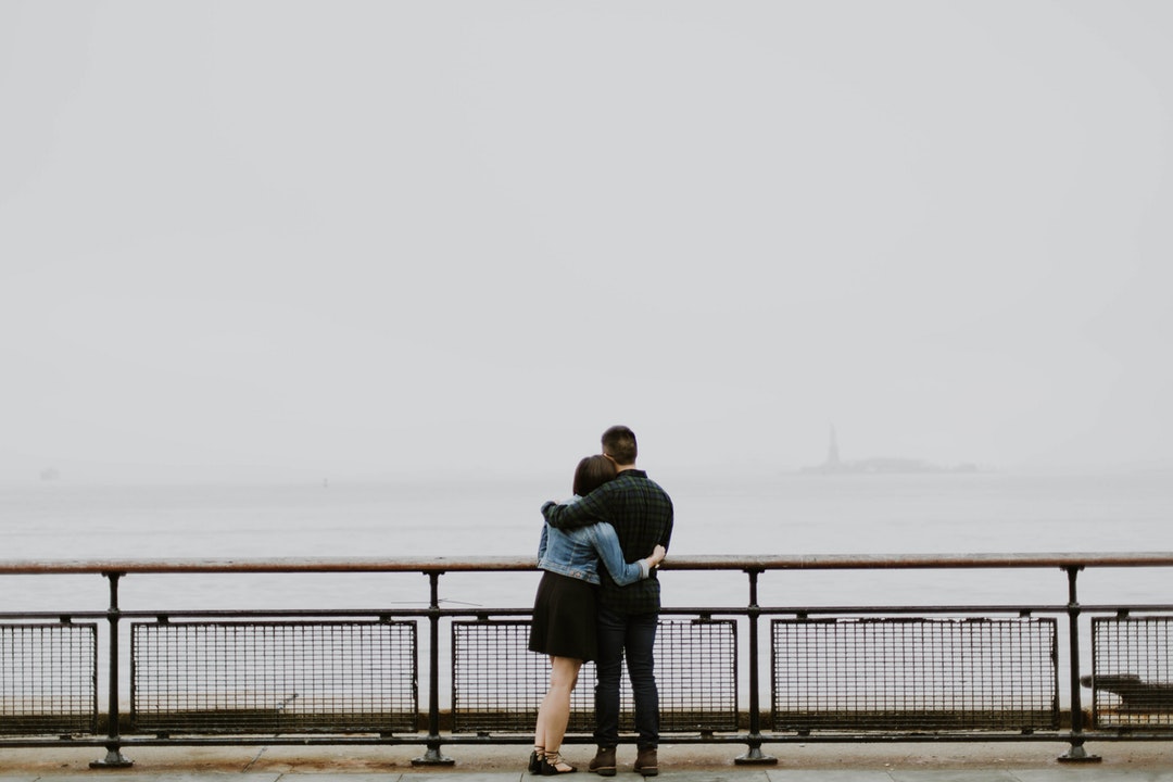 A couple holds one another on a pier overlooking a body of water and overcast sky