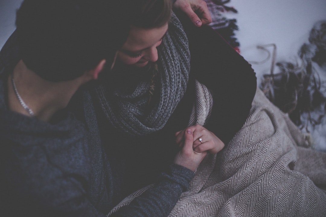 Man and woman snuggle under a blanket, seated and holding hands. She wears an engagement ring.