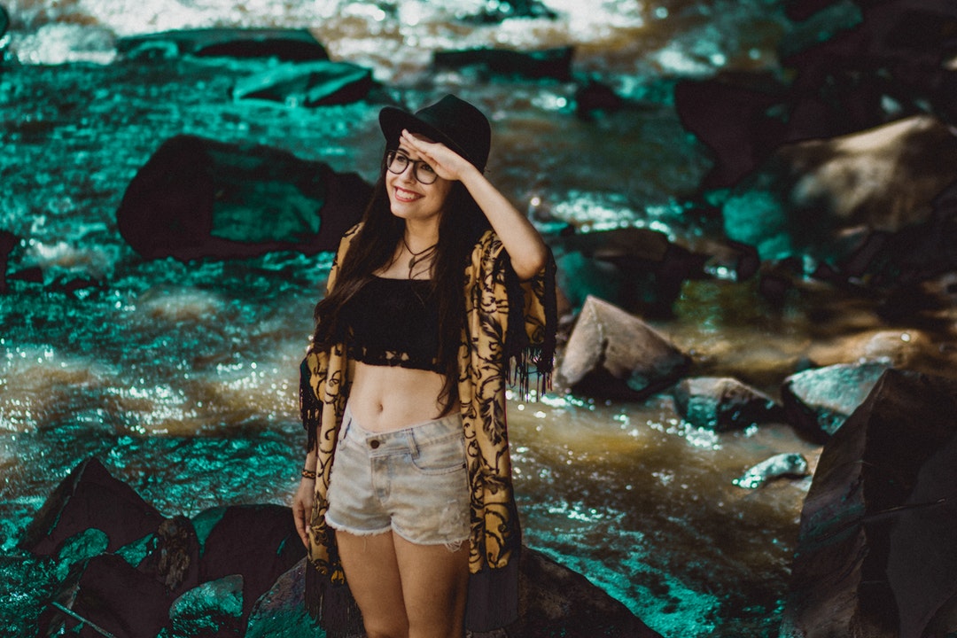 A smiling woman in a hat and glasses shades her eyes aside a blue, rocky stream