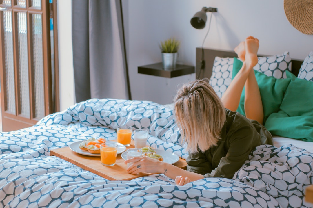 A woman laying on her bed, eating breakfast on a blue and white bedspread in her bedroom in her apartment in Barcelona