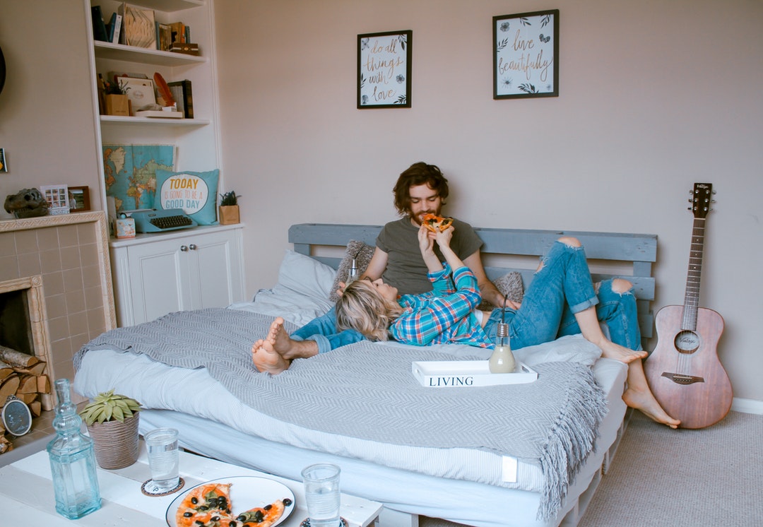Young couple on bed feeding each other pizza with fireplace, guitar and wall art in teddington
