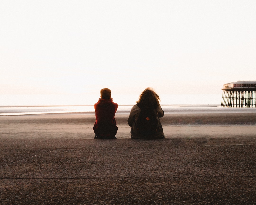 Two people sitting on the sands of Blackpool beach with a pier visible in the distance