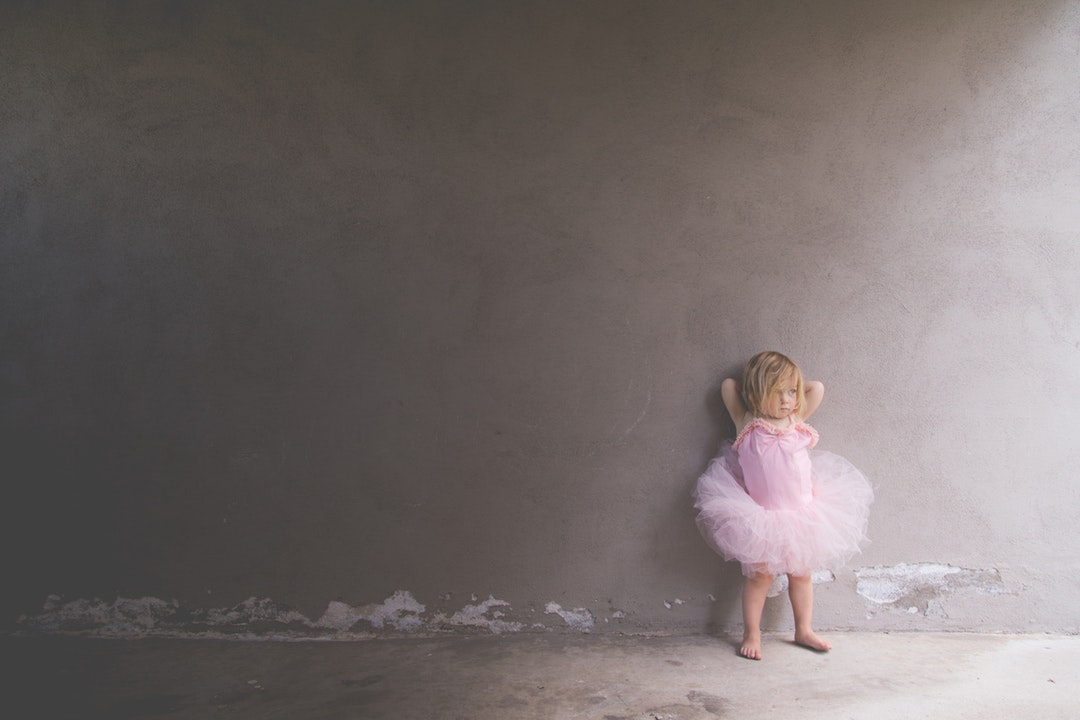 A shadow falls on a child wearing a pink leotard and tutu, leaning against a wall