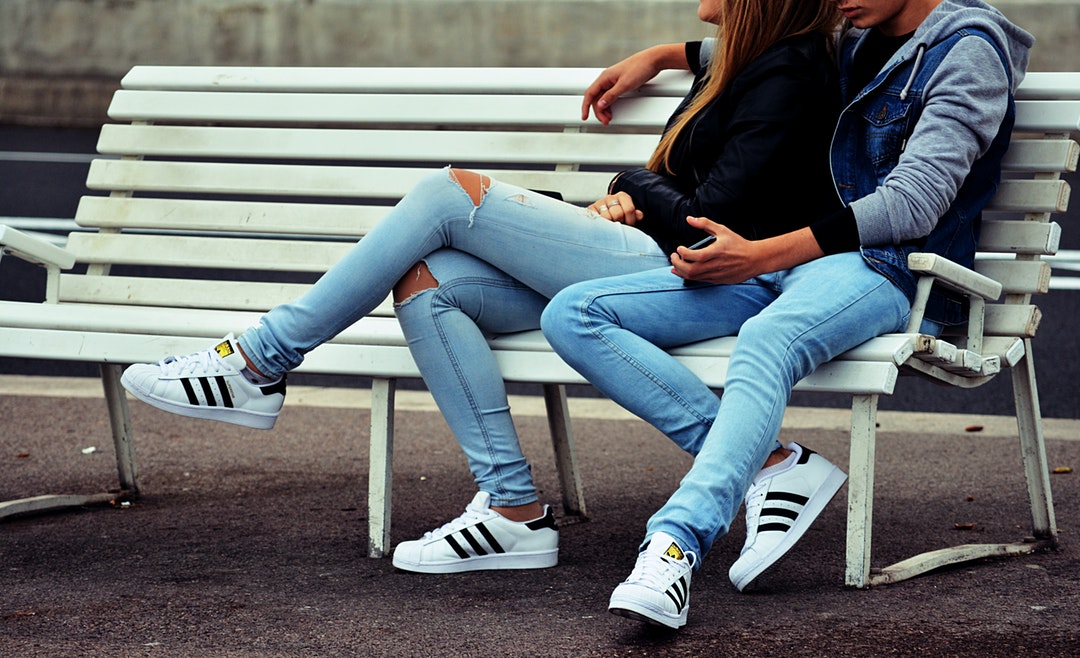 A man puts his hand around a woman in jeans, sitting on a white bench outdoors