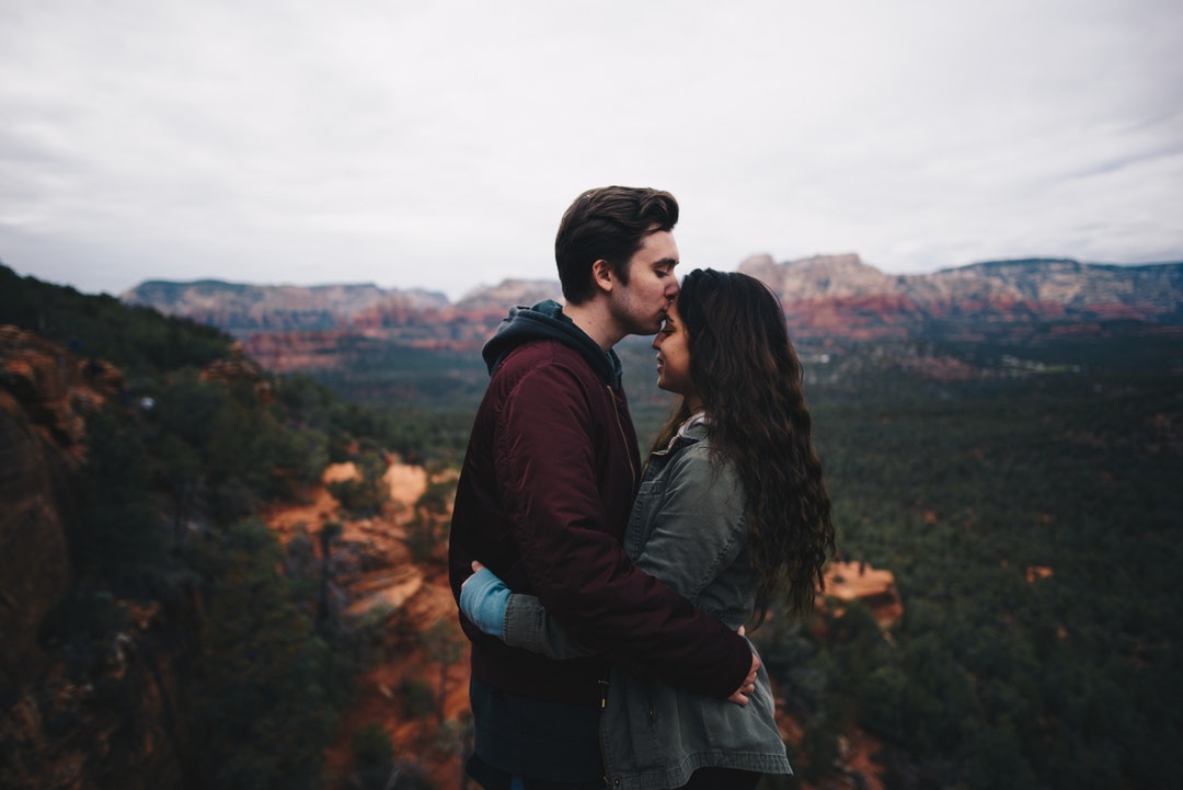 A man kisses a woman's forehead as they embrace in the mountains