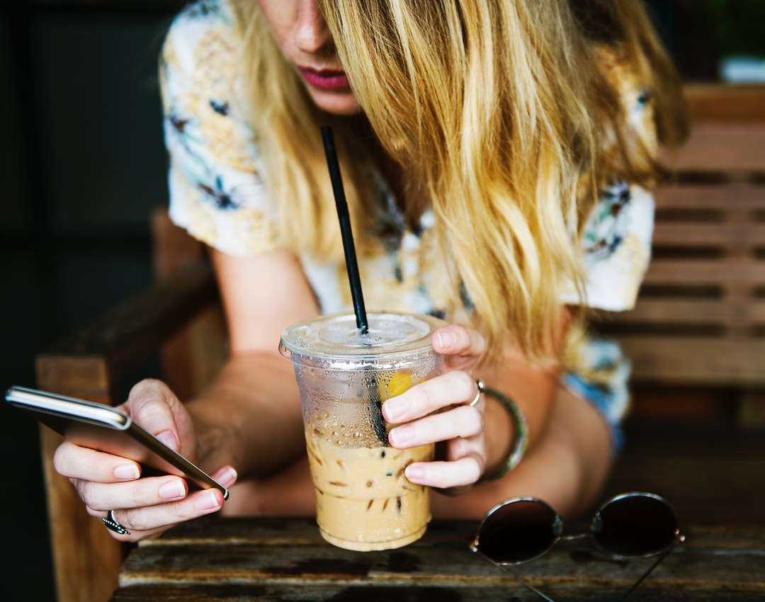 A blonde woman drinking iced coffee while using an iPhone