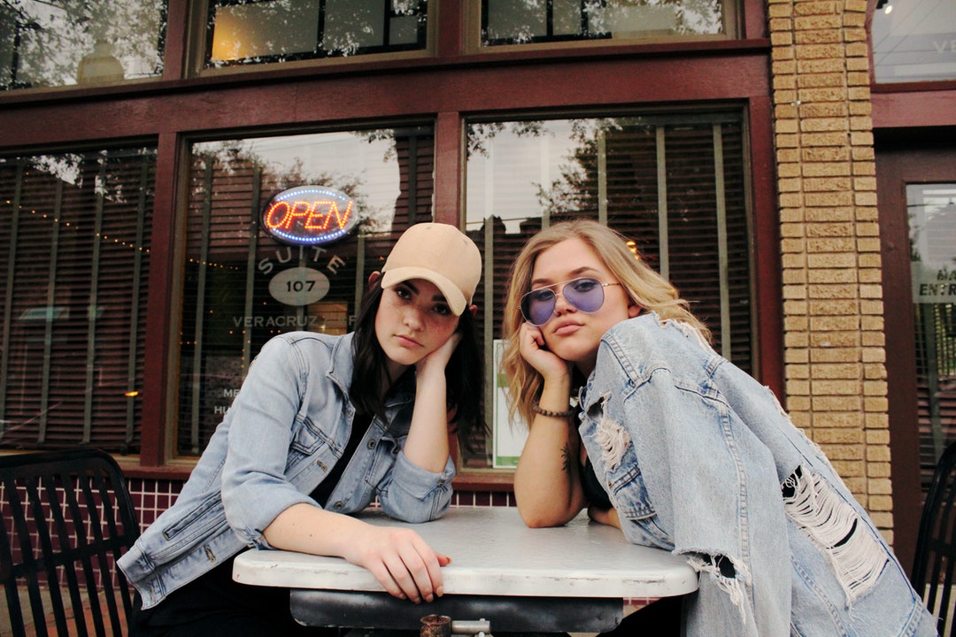 Two women in denim sit at a table in front of a business in Dallas