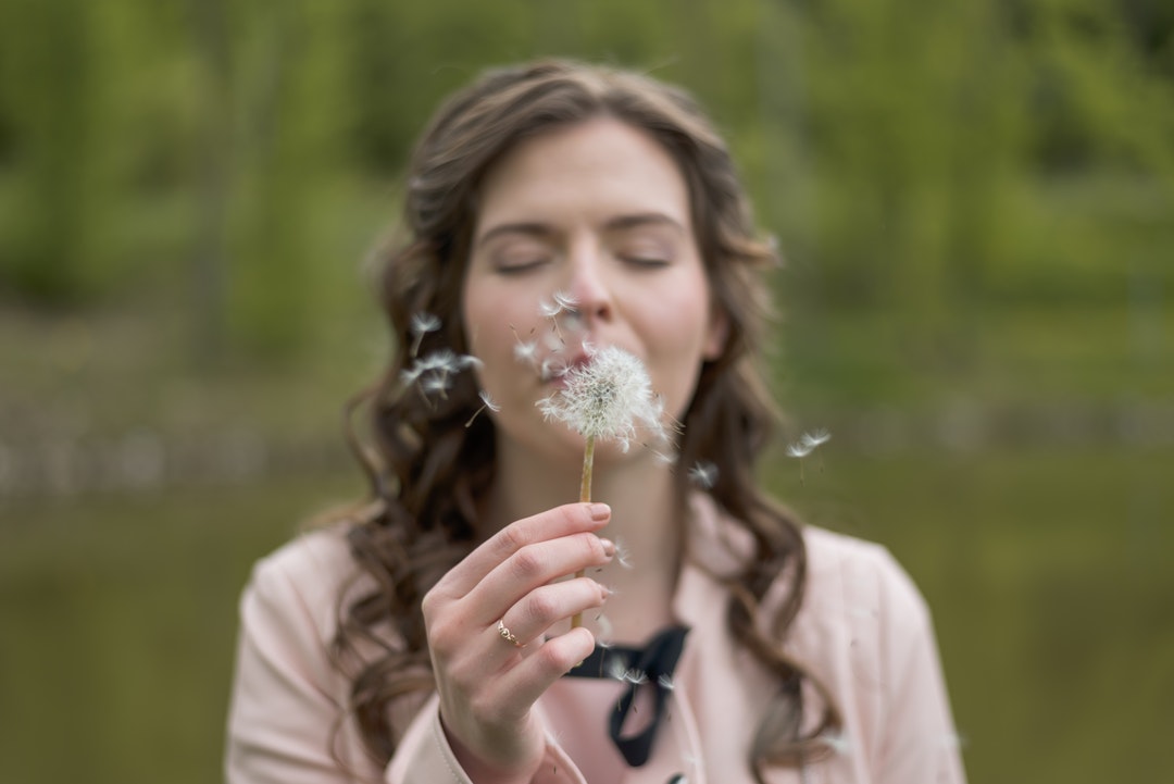 A woman with eyes closed blows seeds from a dandelion against a green background