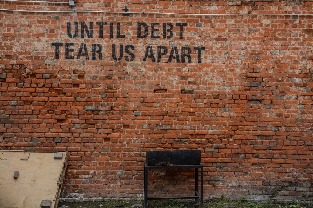 Graffiti on an old brick wall reads “until debt tear us apart”