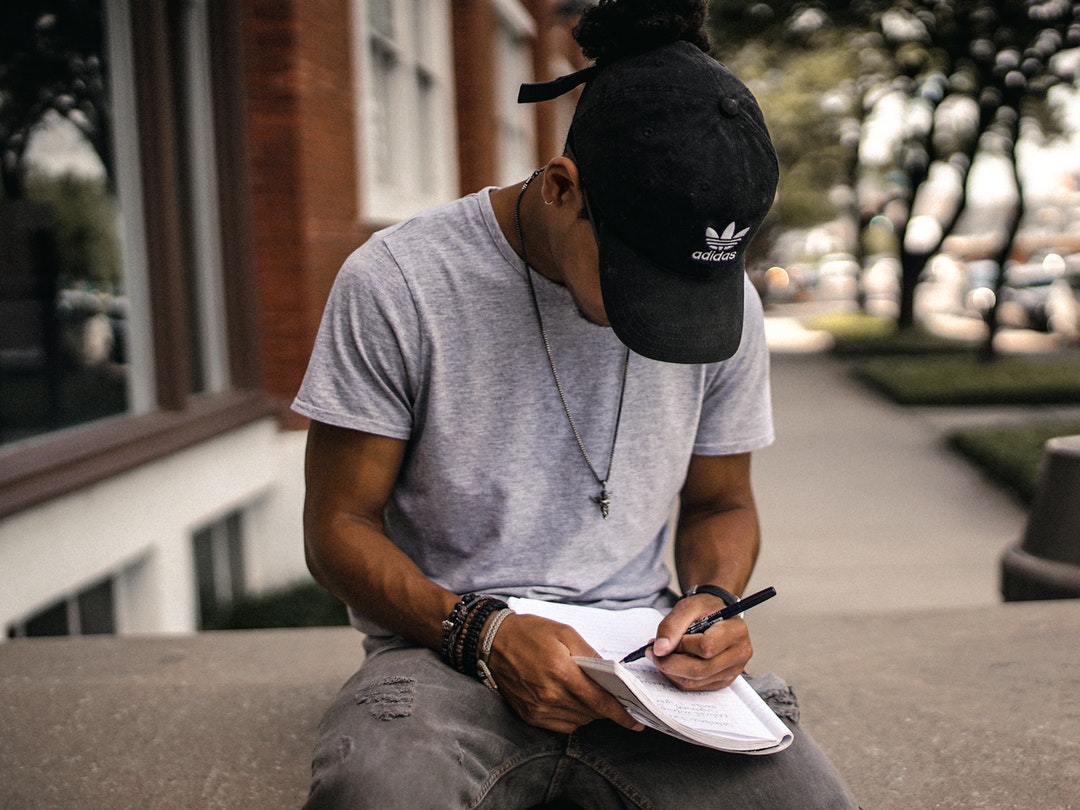 A man in a baseball cap taking notes while sitting on a concrete ledge outdoors