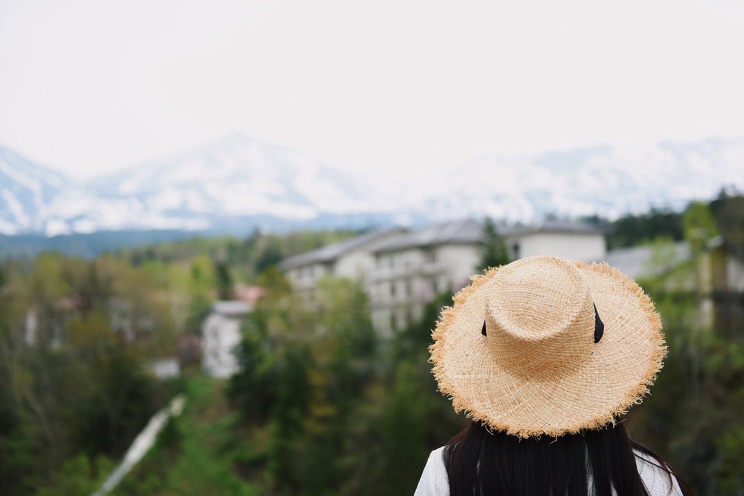 A woman with dark hair wearing a straw hat looking at a village surrounded by trees under a snowy white mountain