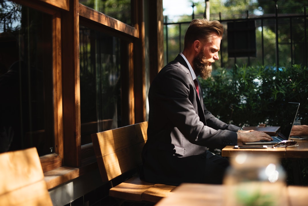 A bearded man wearing a suit sitting outside and working on his laptop at a patio table