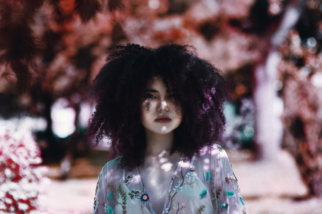 A woman with purple-tinted curly hair in a sheer top stands before a blurry outdoor background