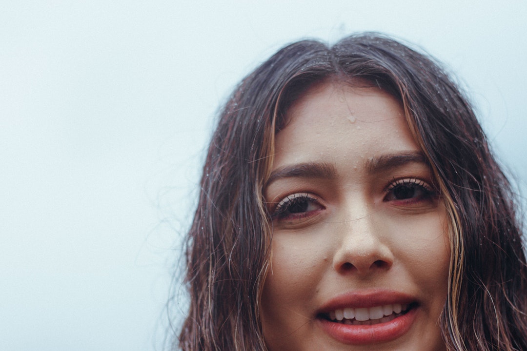 A woman with long brown wet hair and dripping forehead smiles against a blue background