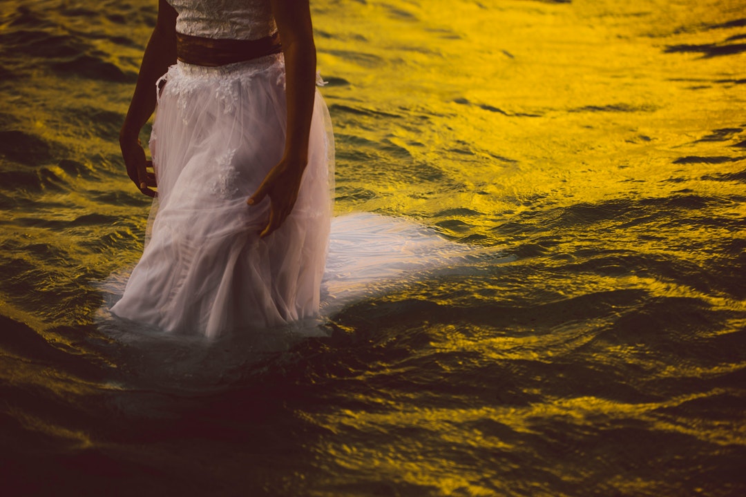 A woman walking in the sea in a white dress, as the water reflects the Rarotonga sunset