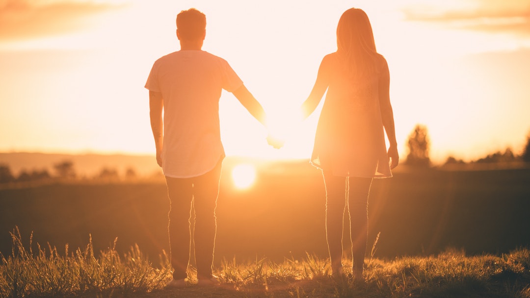 Man and woman holding hands, looking away towards a setting sun