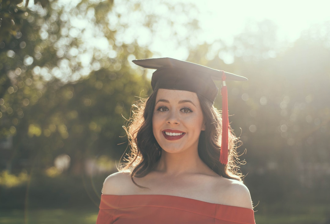 A woman in a red off-shoulder top wearing the mortarboard smiles at the camera.