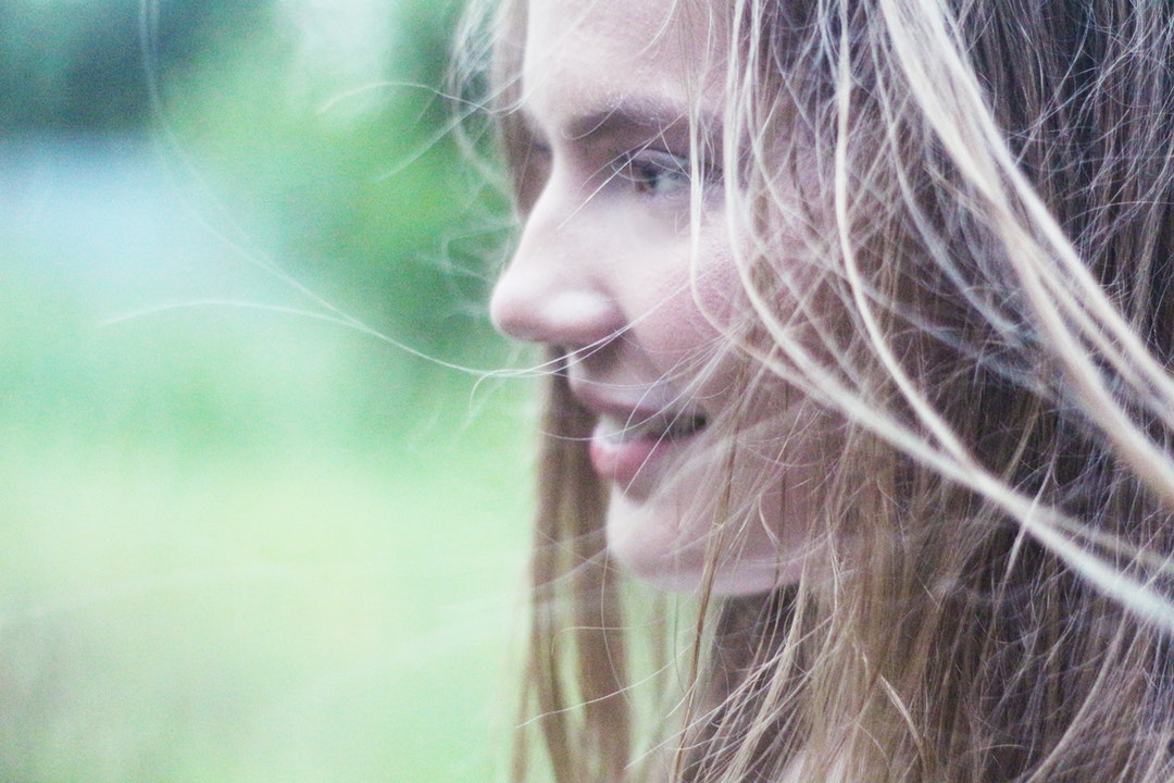 A woman with windswept blonde hair smiles to the side against a blurry green background