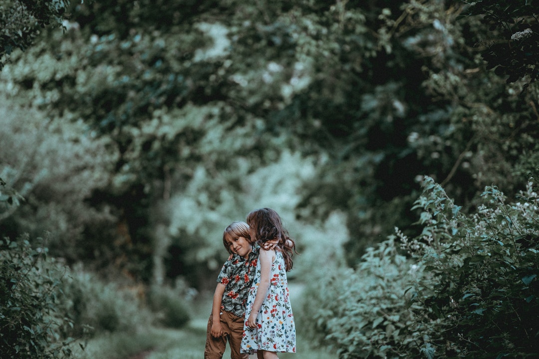Two young children, a boy and girl, hug outdoors in a green forested area