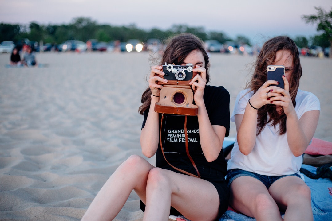 Two female friends are taking photos with a vintage camera and a smartphone at the beach.