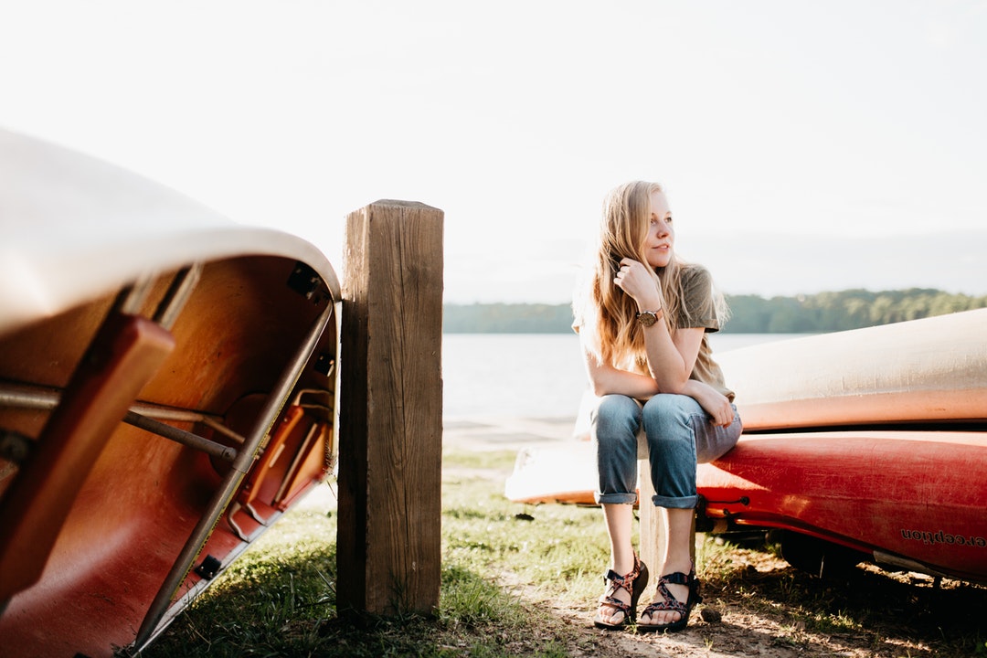 Woman with blonde hair sitting on upside-down red canoe in front of lake and mountains