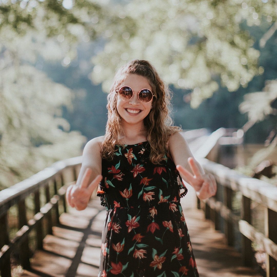 A smiling woman in sunglasses and a dress stands on a pier holding up peace signs