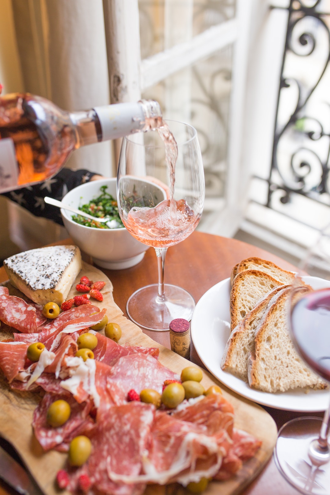 A person pouring rose into a wine glass, a cheese board with brie and salami, and bread on a table in Paris