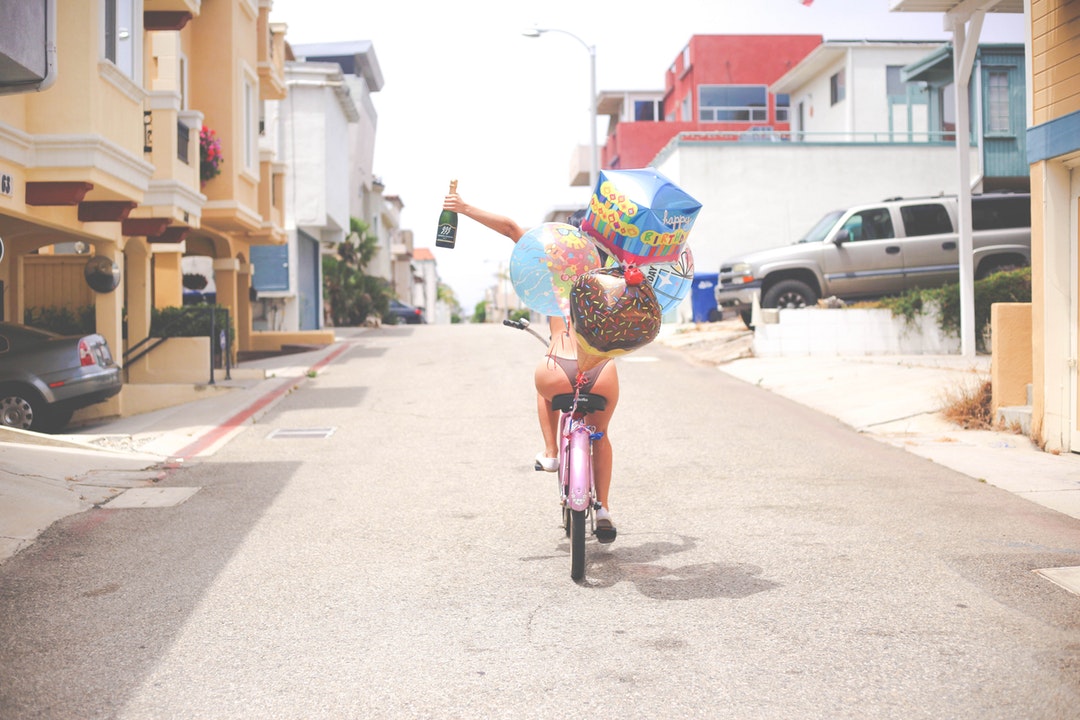 Woman driving a balloon decorated bicycle celebrating and holding a champagne bottle on the street at Hermosa Beach