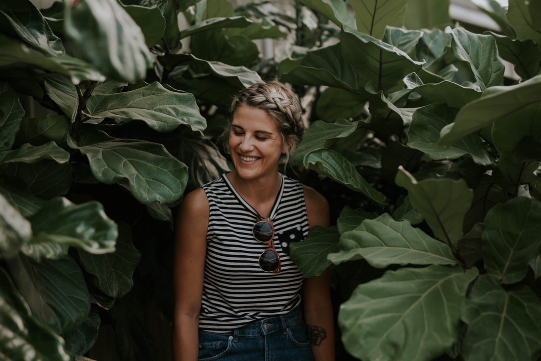 A woman in a tank top smiles with her eyes closed, standing among foliage