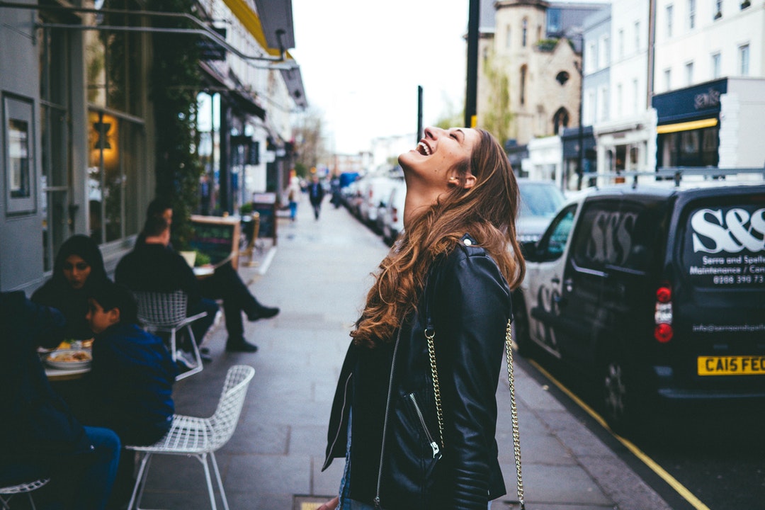 A woman in a leather jacket throws her head back and laughs on a city sidewalk