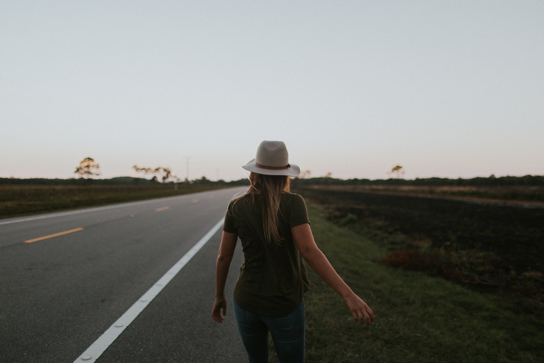 A woman wearing a hat walks away along a highway in daytime sunlight