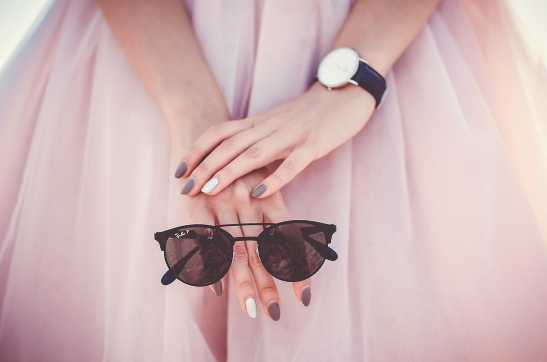 A close-up of a woman's hands with colored nails holding a pair of sunglasses.