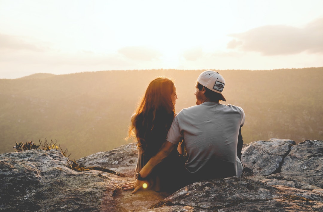 Couple sitting on a rocky mountain looking at each other as sunset colors the sky
