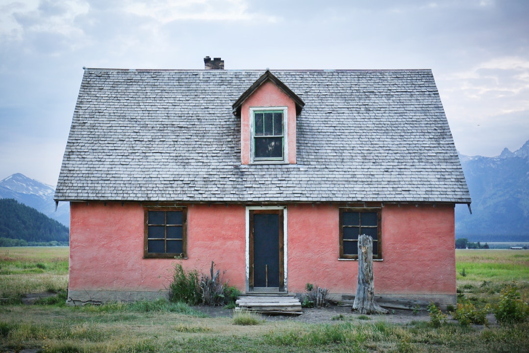 Small pink house with 3 windows in a grassy field with mountains in the background