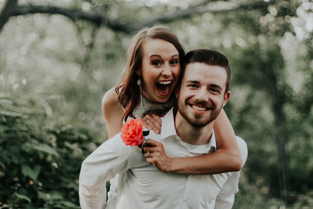 A smiling man gives his fiancee a piggyback ride as she holds a flower