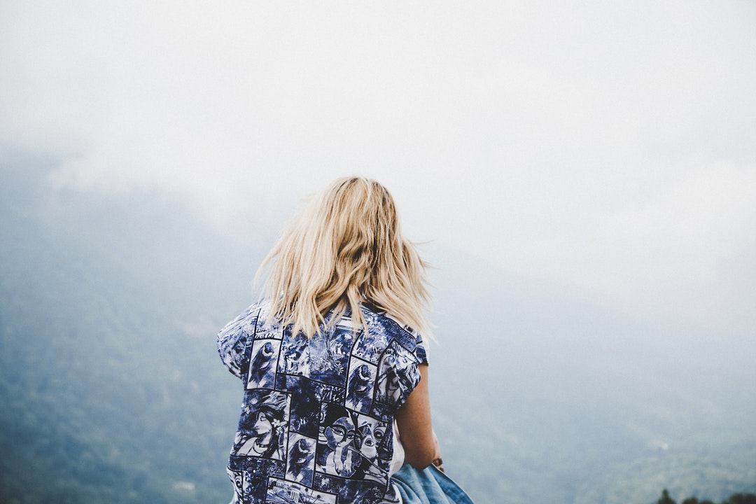 Woman with blonde hair and blue dress standing on a hill, looking at fog covering mountain